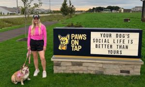 Woman with her bulldog on a leash standing next to the Paws on Tap sign in Whitestown, Indiana