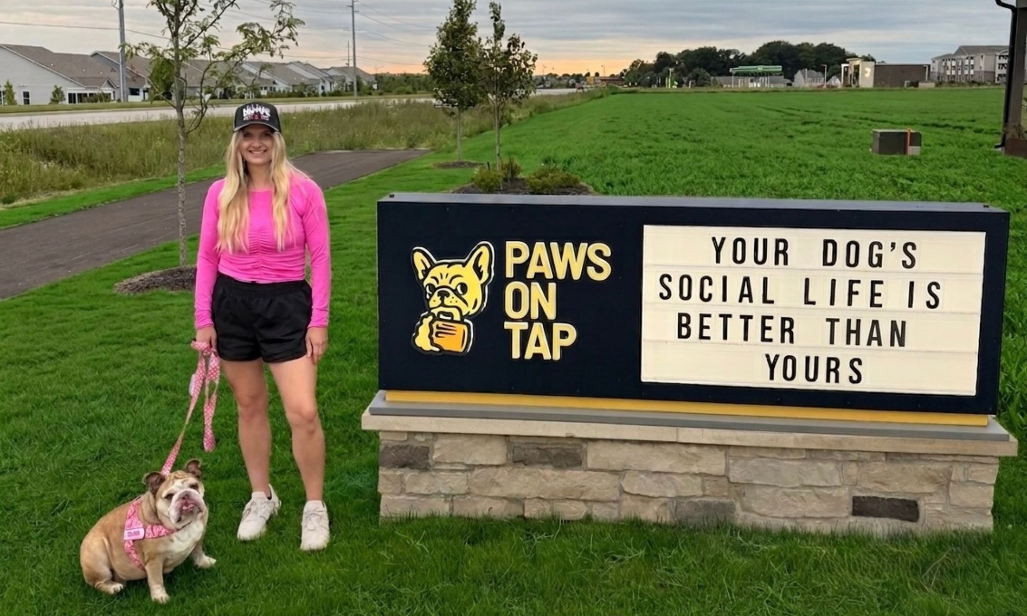 Woman with her bulldog on a leash standing next to the Paws on Tap sign in Whitestown, Indiana